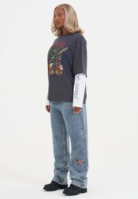 Grijze graphic t-shirt met kleurig drakenontwerp, gedragen over een witte langemouwen shirt. Lichtblauwe jeans met rozenborduursel, zwarte schoenen.