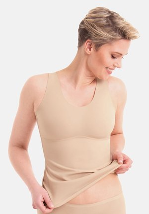 Woman with short hair smiles while pulling up her beige seamless tank top to show its stretch and fit.