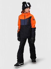 Παιδί ντυμένο με πορτοκαλί και μαύρο μπουφάν σκι, μαύρα παντελόνια, γάντια, σκουφάκι και γυαλιά σκι, που στέκεται με το κεφάλι γυρισμένο στο πλάι.