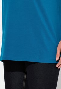 Tyrkysová bavlněná košile s krátkým rukávem, zakřiveným lemem a detaily švů na bočních švech, v kombinaci s tmavými džíny leggingy.