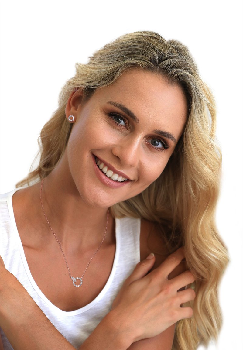 Sølv halskæde med en rund vedhæng, der har klare sten, båret over en hvid ærmeløs top. Modellen har langt bølget blond hår.