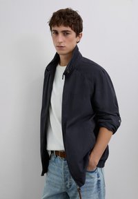 Un jeune homme portant une veste marine avec les manches retroussées, une chemise blanche et un jean bleu clair, se tenant contre un fond gris clair uni.