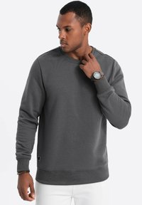 Felpa grigia con scollo rotondo, maniche lunghe e polsini e orlo a costine. Indossata con un orologio da polso. Texture liscia.