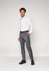 Chemise blanche à manches longues avec boutons, pantalon gris ajusté, ceinture noire et chaussures à lacets noires. Tissu lisse, design ajusté, couleurs neutres.