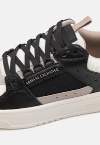 Sneaker nera e beige con finitura testurizzata, dotata di lacci neri, una tomaia in rete bianca e un logo del marchio in rilievo sul lato.