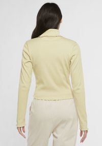 Ljusgul ribbad långärmad topp med scallopkrage och -slut. Har en figurnära design och mjuk textur. Knapppynt på ärmen.