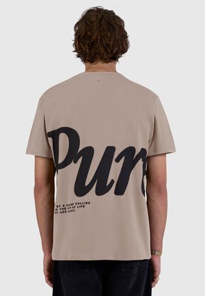 Man with short curly hair wearing beige t-shirt with large black text "Pur" and small text at bottom, shown from the back.