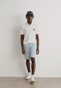 White polo shirt with a red logo, paired with light blue striped shorts. The model wears a dark cap and white sneakers.