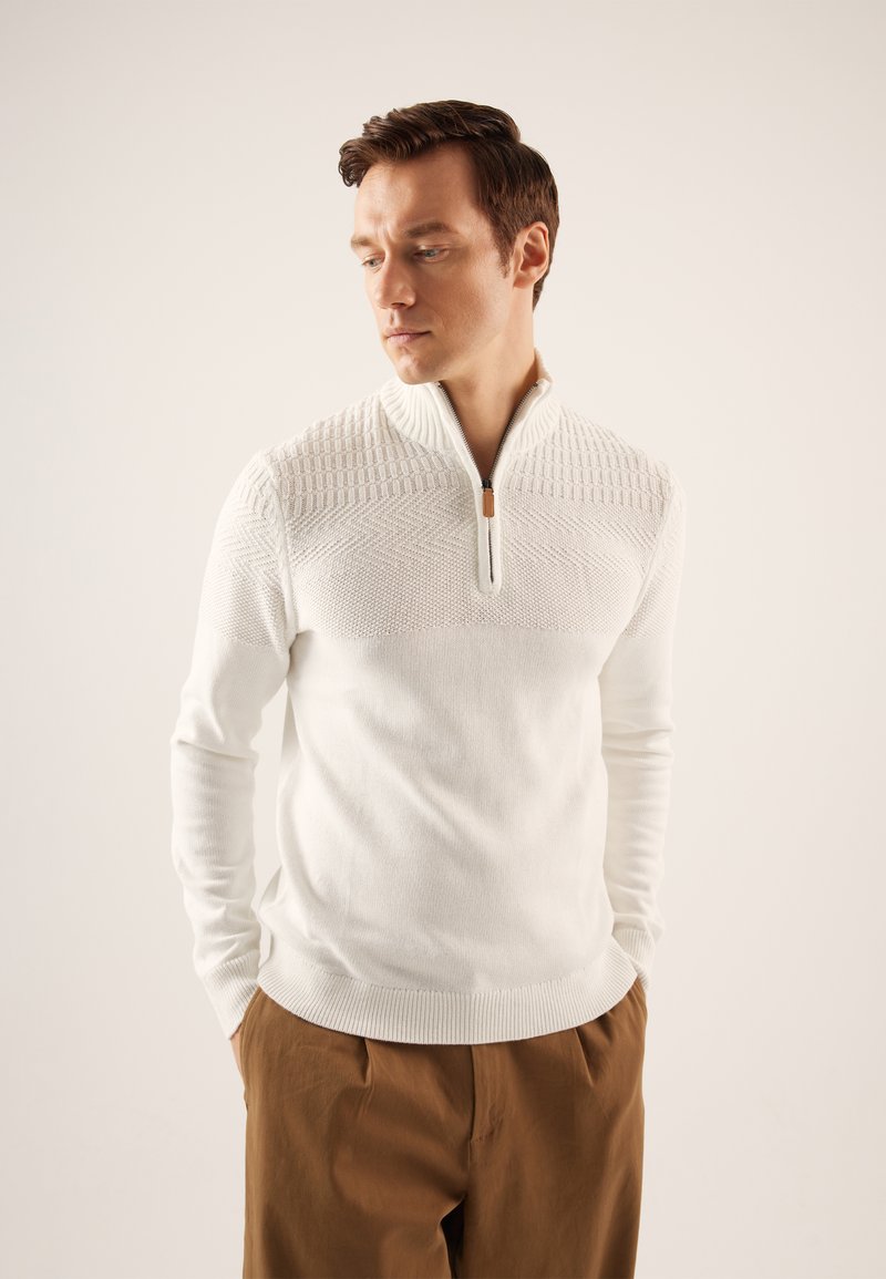 Suéter blanco de punto con cremallera de un cuarto, con un patrón texturizado en los hombros, dobladillo inferior ajustado y puños acanalados. Combinado con pantalones marrones.