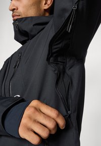 Black outerwear with high collar and multiple zippered pockets. Features a textured, smooth fabric and fitted cuffs for a streamlined look.
