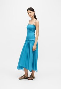 Jeune femme debout portant une robe turquoise sans manches et des sandales plates marron sur un fond clair uni.