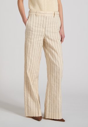 Beżowe szerokie spodnie z cienkimi pionowymi czarnymi paskami, zestawione z brązowymi spiczastymi butami i rękami w kieszeniach.