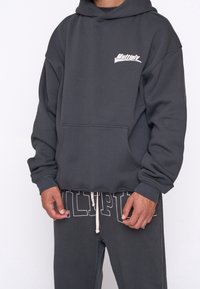 Sudadera gris con un bolsillo tipo canguro y un logo blanco. Combinada con pantalones de chándal grises a juego con lettering en relieve y cordón.