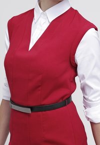 Červené šaty bez rukávů s výstřihem do V a přizpůsobeným střihem, s černým páskem v pase a bílou košilí pod nimi.