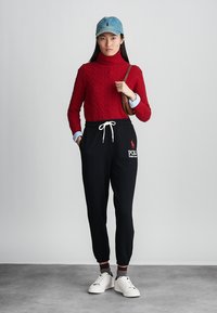 Rød kabelstrikket turtleneck genser, sorte joggebukser med snøring og logo, hvite sneakers, denim caps. Enkelt, uformelt antrekk.