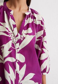 Blouse violette avec un grand motif floral blanc, présentant un décolleté en V, des manches courtes à volants et une fermeture à boutons avec des boutons assortis.