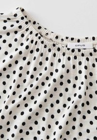 Encolure de chemise blanche à pois noirs et col élastique froncé montrant l'étiquette de la marque OPUS.