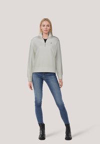 Pullover verde chiaro con zip, maniche a coste e orlo a coste, caratterizzato da un piccolo logo sul petto, abbinato a jeans blue e stivaletti neri.