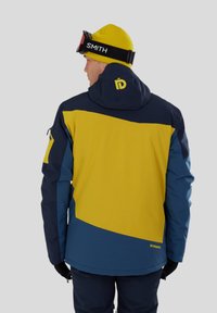 Skijakke i blå, gul og marineblå; har en hætte og logo på ryggen. Struktureret stof med et geometrisk farveblokdesign.