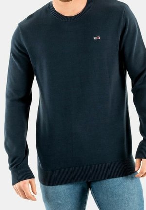 Man wearing a dark navy blue ribbed crewneck sweater with a small rectangular logo on the chest and light blue jeans.
