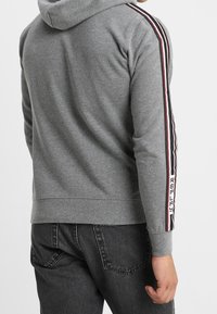 Felpa grigia con tessuto strutturato, caratterizzata da strisce a contrasto in rosso e nero lungo le maniche e un logo sul braccio sinistro.