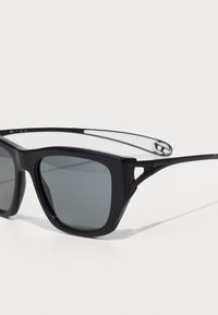 Óculos de sol pretos com lentes retangulares, apresentando um acabamento mate, hastes finas e um design distintivo com recorte próximo às dobradiças.