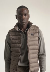 Chaleco acolchado marrón con cuello alto, cierre de cremallera y parche con logo, llevado sobre una camisa de manga larga gris. Textura suave y patrón acolchado.