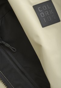 Interno della giacca nero e beige con una tasca con zip, materiale liscio e un'etichetta nera testurizzata con testo in rilievo.