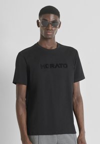 Camiseta de algodón negra con un logo en relieve en el pecho. Cuello redondo simple y mangas cortas. Modelo usando gafas de sol oscuras.