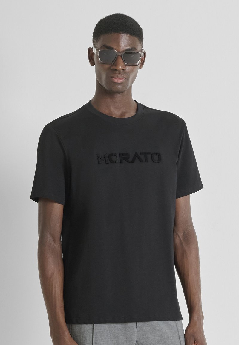 Camiseta de algodón negra con un logo en relieve en el pecho. Cuello redondo simple y mangas cortas. Modelo usando gafas de sol oscuras.