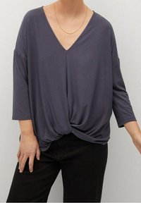 Top grigio largo con scollatura a V profonda, maniche a tre quarti e dettaglio annodato sul davanti. Realizzato in un tessuto morbido e leggero.