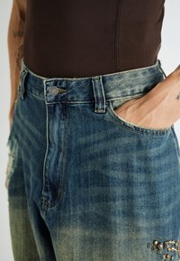 Pantalones vaqueros de mezclilla azul de talle alto con textura desgastada, detalles desgarrados y tachuelas decorativas en el bolsillo delantero.