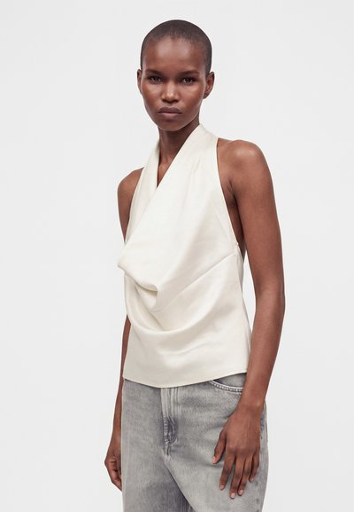 Femme à la tête rasée portant un haut blanc sans manches drapé et un jean gris clair, debout devant un fond clair uni.