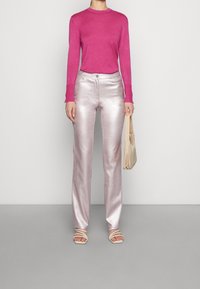 Fialová dlouhá košile, lesklé světle růžové široké kalhoty a béžové sandály na řemínku. Model drží béžovou tašku a stojí na neutrálním pozadí.