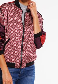 Red and black patterned bomber jacket with diamond shapes, zip closure, ribbed cuffs, and textured sleeves. Worn over a gray top.