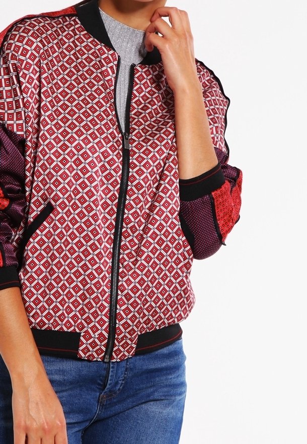 Red and black patterned bomber jacket with diamond shapes, zip closure, ribbed cuffs, and textured sleeves. Worn over a gray top.