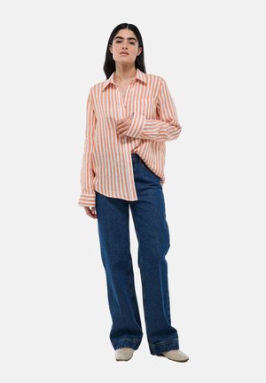 Chemise à boutons rayée orange et blanche, assortie à un jean bleu large. La chemise a un col et une seule poche poitrine.