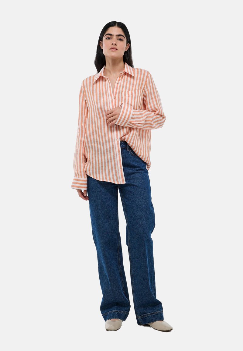 Chemise à boutons rayée orange et blanche, assortie à un jean bleu large. La chemise a un col et une seule poche poitrine.