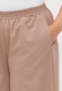 Beige, lightweight trousers with an elastic waistband and side pockets. The fabric has a soft texture and a slightly flowing design.