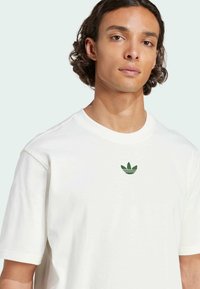 White cotton t-shirt with crew neck, featuring a small green Adidas logo on the chest. Short sleeves and a relaxed fit.