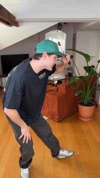 Žalia kepurė, juoda perdidinta marškinėliai su logotipu, tamsios kelnės ir sidabrinės sportbačiai. Stovi viduje šalia vazoninės gėlės ir oranžinės sofos.