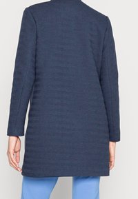 Marinblå kappa med fiskbensmönster, långa ärmar och rak skärning. Har en ren halsringning och textil med struktur.