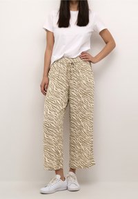 Femme portant un t-shirt blanc, un pantalon large à rayures zèbres beige et blanc, et des baskets blanches, se tenant avec une main sur la hanche devant un fond uni.