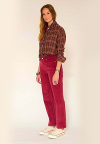 Chemise en flanelle à carreaux dans les tons violet et vert, portée décontractée. Pantalons en velours côtelé bordeaux avec poches avant. Baskets beiges avec des accents rouges.