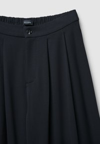 Marineblaue Hosen mit einem Bund, der über Gummi und einen Knopfverschluss verfügt. Enthält vordere Falten und eine Seitentasche. Glatte Textur, taillierte Passform.