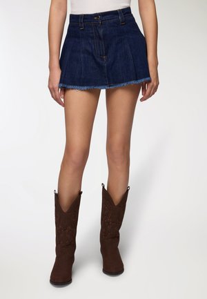 Gonna mini in denim blu scuro con orlo sfilacciato, dotata di chiusura frontale con bottoni e due tasche laterali, abbinata a stivali da cowboy marroni.