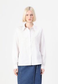Camicia bianca con bottoni e design floreale ricamato sul petto, maniche lunghe e colletto; indossata con una gonna di jeans.