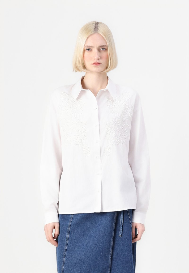 Camicia bianca con bottoni e design floreale ricamato sul petto, maniche lunghe e colletto; indossata con una gonna di jeans.