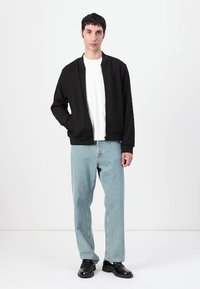 Svart bomberjakke med glidelås, brukt over en hvit t-skjorte, kombinert med lyseblå jeans og svarte sko. Enkel, moderne design.