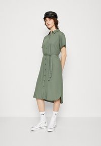 Abito camicia verde oliva con maniche corte, apertura frontale con bottoni, vita annodata e orlo curvo. Abbinato a sneakers bianche e calzini alla caviglia.